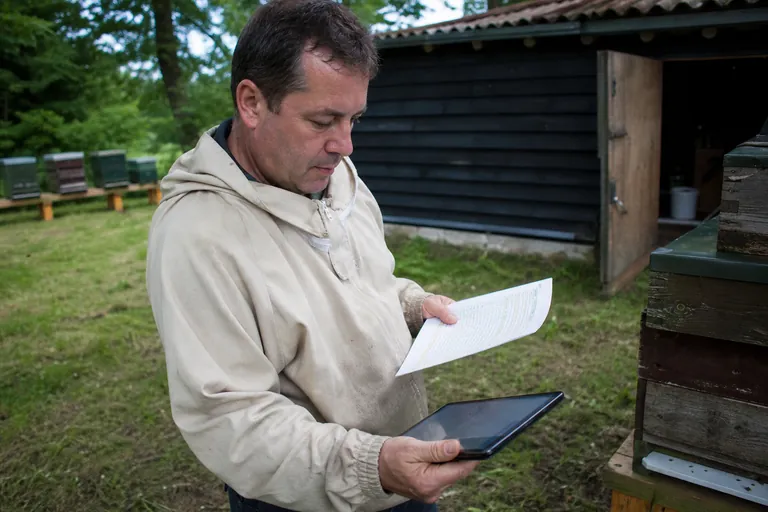 Imker hält eine Papierstockkarte und betrachtet eine Völkerübersicht auf einem Tablet